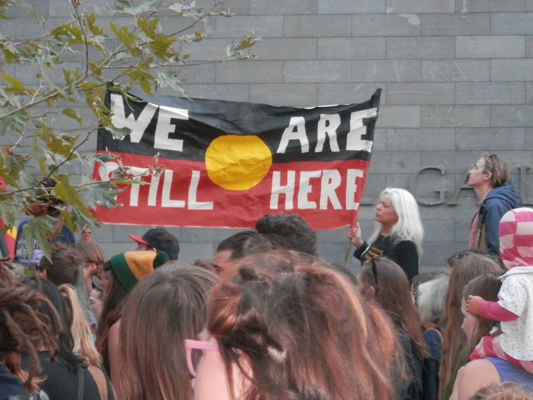 Protest banner reads "We Are Still Here" on Aboriginal flag.