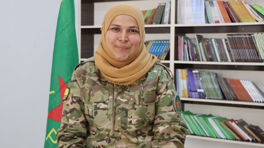 Woman wearing a hijab and military uniform sitting in front of a bookshelf