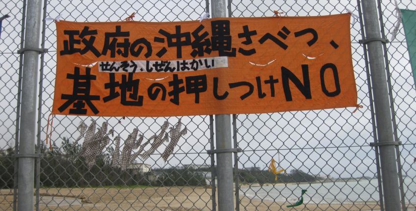 An orange banner with Japanese text hangs on a chain-link fence overlooking an Okinawan beach, protesting the government and the forcing of military bases onto the region.