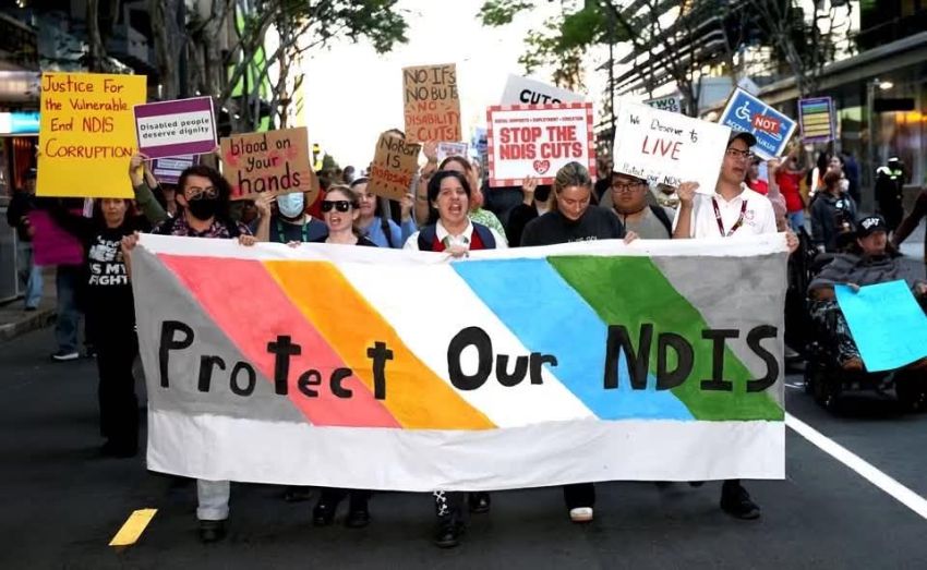Protestors marching and holding a banner that reads Protect our NDIS at at rally against cuts to the NDIS