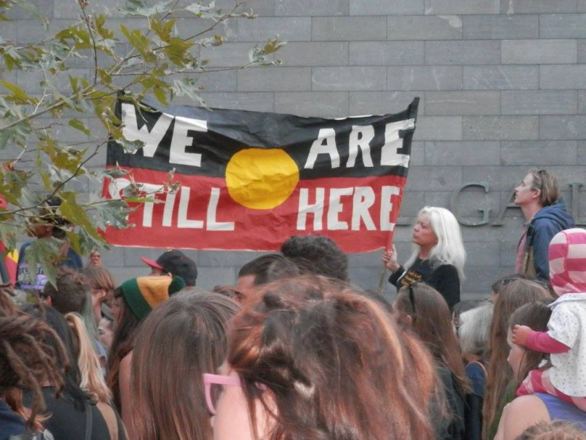 Protest banner reads "We Are Still Here" on Aboriginal flag.
