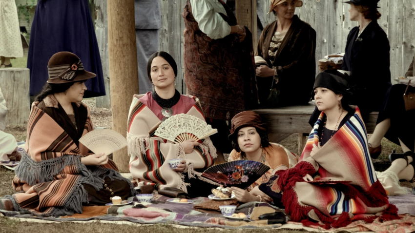 Still from the film, Killers of the Flower Moon, showing Mollie sitting with her sisters on a large blanket on the ground. All of them are holding fans.
