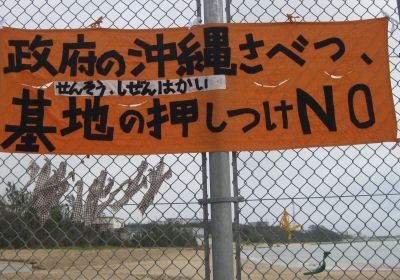 An orange banner with Japanese text hangs on a chain-link fence overlooking an Okinawan beach, protesting the government and the forcing of military bases onto the region.
