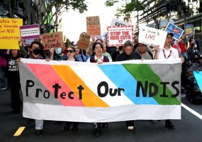 Protestors marching and holding a banner that reads Protect our NDIS at at rally against cuts to the NDIS